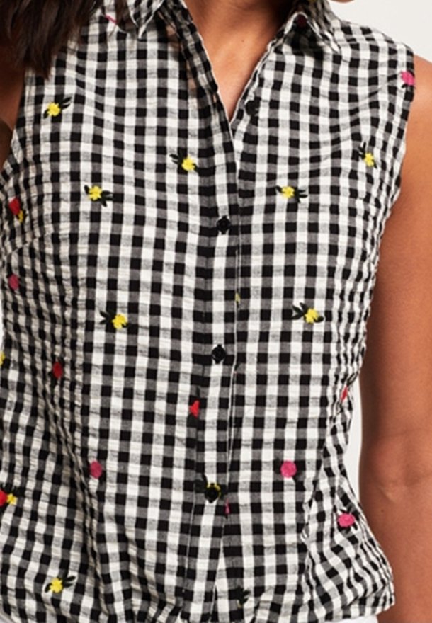 Femme portant une chemise à carreaux noir et blanc sans manches, avec un col et de petites fleurs brodées jaunes et roses. Seul le haut du torse est visible.