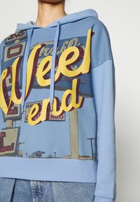 Niebieska bluza z kapturem z dużą żółtą grafiką "Weekend". Charakteryzuje się nadrukiem vintage w stylu znaku. Miękka tkanina, regulowany sznurek i ściągacz na hemie.