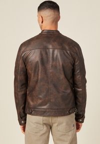 Man met kort haar en een hoepeloorbel, gekleed in een donkerbruine leren jas en lichtbeige broek, met de rug naar de camera toe.