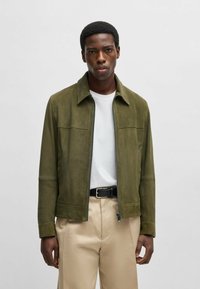 Olivengrön mockajacka med dragkedja fram, spetsig krage och två bröstfickor. Bärs över en vit t-shirt och beige byxor.