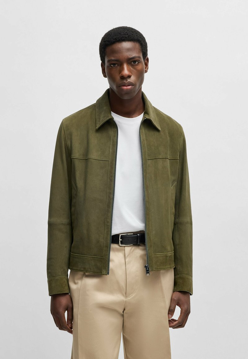 Olivengrön mockajacka med dragkedja fram, spetsig krage och två bröstfickor. Bärs över en vit t-shirt och beige byxor.