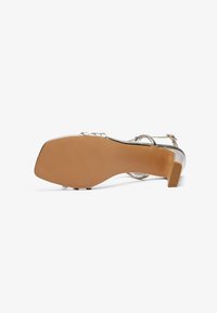 Heeled sandal with a metallic silver finish, open design, and an ankle strap. Square toe and beige rubber sole.