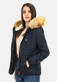 Vrouw in een marineblauwe parka met een met bont gevoerde capuchon, een beige shirt en blauwe spijkerbroek, staand tegen een effen lichte achtergrond.