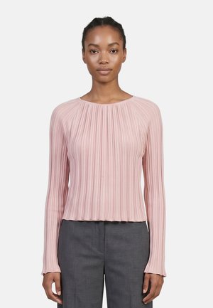 Femme portant un haut côtelé à manches longues rose clair et un pantalon gris, debout devant un fond blanc uni.