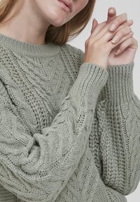 Grüner Strickpullover mit Zopfmuster, ribbeliichem Rundhalsausschnitt und strukturierten Ärmeln. Weiches Material mit einer gemütlichen Erscheinung.