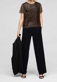 Top in paillettes marroni con maniche corte trasparenti, abbinato a pantaloni larghi blu navy. Il modello tiene un blazer nero e indossa sandali neri.