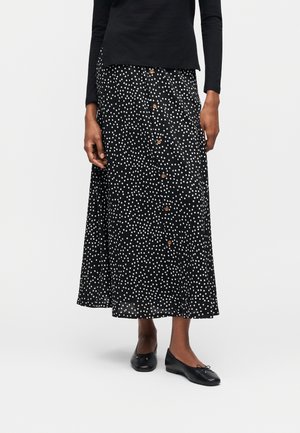 Γυναίκα που φοράει μαύρη midi φούστα με λευκές πουά και κουμπιά στο μπροστινό μέρος, συνδυασμένη με μαύρα μπαλαρίνα παπούτσια και μαύρο τοπ με μακριά μανίκια.