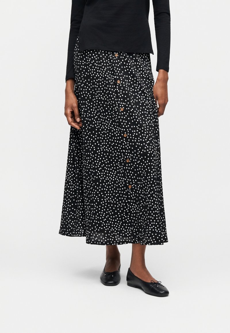 Γυναίκα που φοράει μαύρη midi φούστα με λευκές πουά και κουμπιά στο μπροστινό μέρος, συνδυασμένη με μαύρα μπαλαρίνα παπούτσια και μαύρο τοπ με μακριά μανίκια.