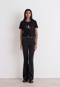 Czarny t-shirt z złotym logo "CK", połączony z czarnymi, wysokim stanem dżinsami w stylu dzwonów. Modelka stoi na tle białej ściany. Czarne buty.
