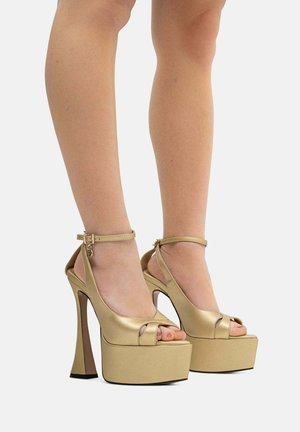 Gambe che indossano sandali con plateau dorati lucidi, tacco alto e cinghietti alla caviglia su uno sfondo chiaro.