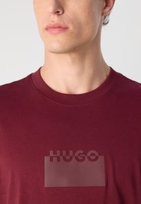 Bordo kolorovo apvalaus kaklo marškinėliai, pagaminti iš minkštos medžiagos, su pusiau permatomu „HUGO“ logotipo pamušalu priekyje. Lygus audinys, paprastas dizainas.