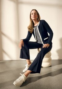 Cardigan marine raccourci avec bord blanc sur un haut à motif, associé à un pantalon large bleu marine et des baskets blanches, assis sur un tabouret sculptural.