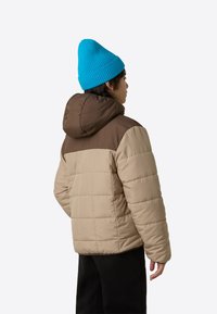 Podložena jakna v bež in rjavi barvi z ruto in navpičnim prešitim vzorcem, nošena z modro pleteno kapo. Vidne so črne hlače.