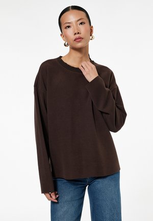 Frau mit dunklem Haar in einem niedrigen Pferdeschwanz trägt goldene Creolen, einen lockeren dunkelbraunen Pullover und blaue Jeans vor einem schlichten weißen Hintergrund.