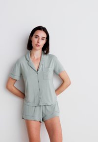 Lichtgroene pyjama-set met korte mouwen, met een knoopsluiting aan de bovenkant en een borstzak, en bijpassende shorts, gemaakt van een zachte, gladde stof.