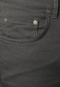 Grijze denim met een lichte structuur, voorzien van een voorkantzak, zilveren metalen klinknagels en contrasterende witte stiksels langs de naden.