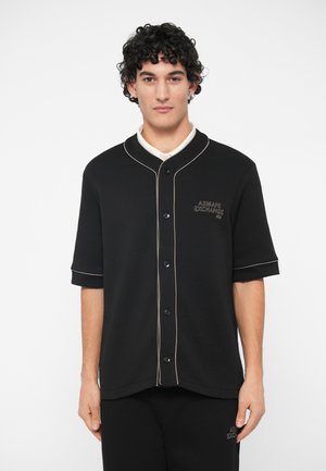 Man med lockigt hår som bär en svart kortärmad skjorta med knappar och vit piping samt Armani Exchange-logotyp, stående mot vit bakgrund.