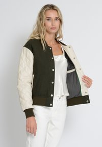 Grøn og creme bomberjakke med kviltede ærmer, trykknaplukning og kontrastfor. Båret over en let satinbluse.