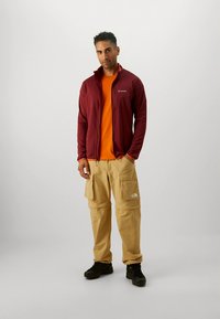 Tumši sarkana rāvējslēdzēja jaka no flīsa virs oranžas T-krekla, kombinēta ar khaki krāsas kargo biksēm ar kabatām. Melni apavi papildina izskatu.