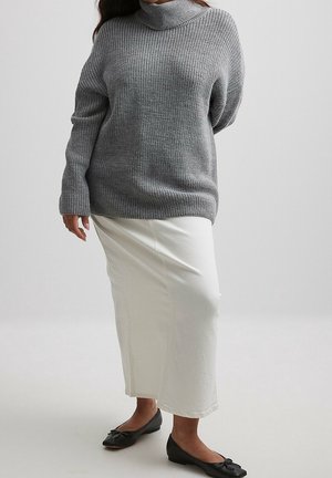 Personne portant un pull à col roulé gris en maille, une longue jupe blanche et des ballerines noires, debout devant un fond gris uni.