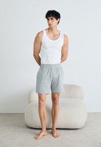 Hvit bomulls tank topp med en liten rød logo, kombinert med grå shorts. Shortsene har elastisk midje og sidelommer. Barfot står på teppe.