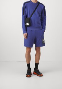 Uomo che indossa un felpa a maniche lunghe blu e pantaloni corti coordinati, con calze nere, sneaker nere e arancioni, e una piccola borsa a tracolla nera.