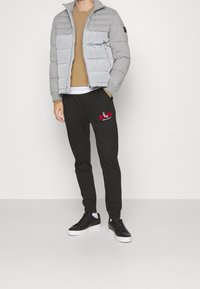 Graue Pufferjacke mit Taschen, brauner Pullover darunter, schwarze Sweatpants mit rotem Logo und schwarze Sneakers mit weißen Sohlen.