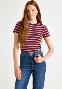 Jonge vrouw met lang, rood haar die een rood, wit en zwart gestreept shirt met korte mouwen en hoge taille blauwe spijkerbroek draagt, tegen een witte achtergrond.