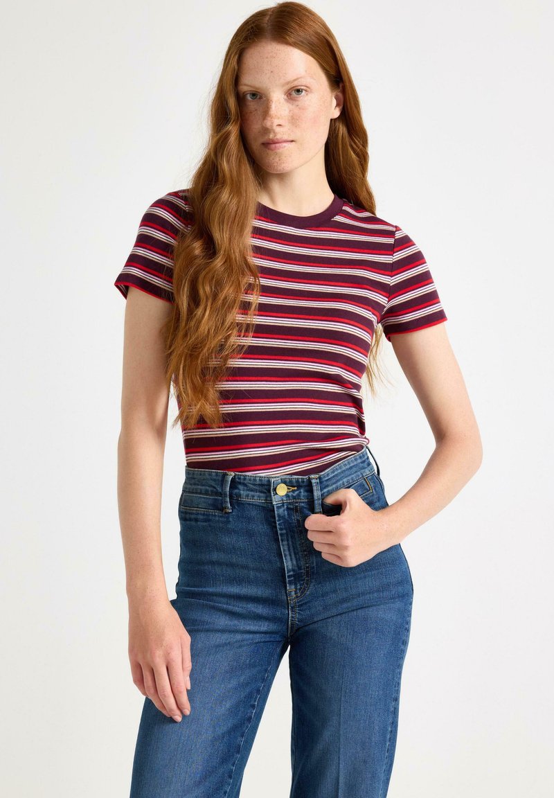 Jonge vrouw met lang, rood haar die een rood, wit en zwart gestreept shirt met korte mouwen en hoge taille blauwe spijkerbroek draagt, tegen een witte achtergrond.