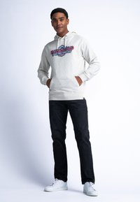Sudadera gris claro con un gráfico de "PETROLIND" en el frente, combinada con jeans negros y zapatillas blancas. Incluye cordones y un bolsillo canguro.