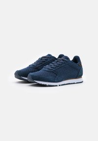 Chaussures de sport bleu marine avec un mélange de tissu texturé et de daim. Fermeture à lacets, languette rembourrée et semelle en caoutchouc blanc avec des accents mouchetés.