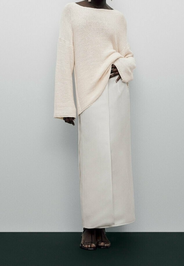 Femme portant un pull tricoté ample crème et une longue jupe crème avec des talons noirs à bout ouvert, sur un fond gris clair uni.