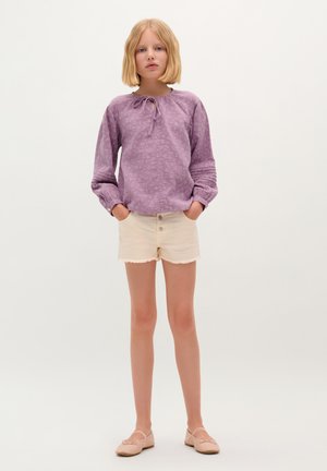 Blusa lunga viola con maniche lunghe, nodo a farfalla e motivo testurizzato, abbinata a pantaloni corti crema con orli sfilacciati e bottoni frontali, indossata con scarpe flat nudo.