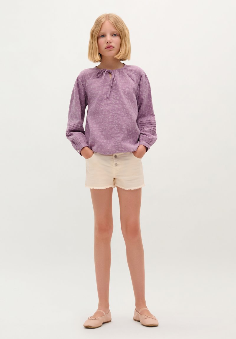 Blusa lunga viola con maniche lunghe, nodo a farfalla e motivo testurizzato, abbinata a pantaloni corti crema con orli sfilacciati e bottoni frontali, indossata con scarpe flat nudo.