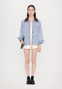 Lichtblauwe denimjack met een bruine leren kraag, oversized pasvorm, twee voorkruiszakken, gedragen over een crèmekleurige playsuit en gestyled met zwarte instappers.