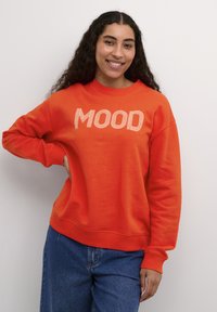 Pomarańczowa bluza z nadrukiem "MOOD" w białym kolorze, z okrągłym dekoltem, ściągaczami przy rękawach i lekko oversize'owym krojem. Noszona z niebieskimi dżinsami.
