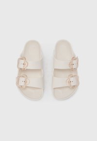 Ανοιχτό μπεζ σανδάλια από υλικό με υφή, με δύο φαρδιές λουρίδες με διαφανή αγκράφες και δ ιαμορφωμένη σόλα.