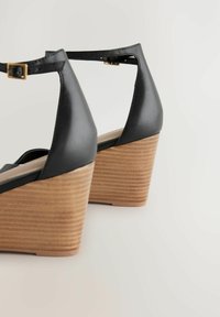 Mustat nahka wedges-kengät, joissa on teksturoitu puinen korko, nilkkaremmillä ja pyöreällä varvastyylillä.