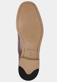 Semelle de chaussure en cuir marron avec une surface lisse beige, une couture visible et un embout en caoutchouc noir. Logo Clarks embossé près du talon.