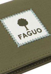 Quadratischer weißer Stofffleck mit blauer Zickzacknaht, einer grünen Baumgrafik und dem Wort "FAGUO" auf olivgrünem Textilhintergrund.