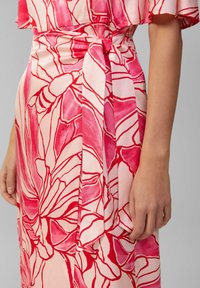 Rosa Blumenkleid mit kräftigen roten Konturen, aus weichem Stoff gefertigt. Verfügt über eine Krawatte in der Taille und eine fließende Silhouette mit kurzen Ärmeln.