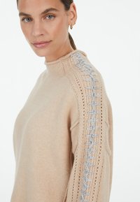 Beige knitted sweater with a high neckline, featuring a textured gray accent along the shoulder. Soft material and relaxed fit.