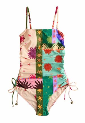 Vrouw staat op het strand met een strapless, kleurrijk badpak met zon- en sterrenpatronen, en houdt een gevlochten tas op haar linkerheup.