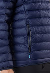 Marineblaue Steppjacke mit quiltierter Textur, mit Reißverschlusstasche und einem dekorativen schwedischen Flaggenakzent nahe dem Saum. Glattes, leichtes Material.