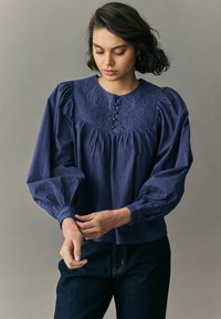 Blouse en coton bleu marine avec un détail brodé au niveau du col, des manches longues bouffantes et une fermeture à boutons. Portée avec un jean foncé.
