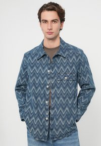 Jachetă denim cu un model zigzag în tonuri de albastru, având închidere cu nasturi pe față, guler ascuțit și un buzunar la piept.