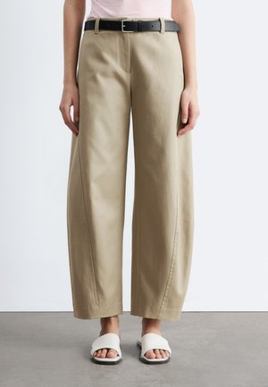 Kobieta w beżowych szerokich spodniach z czarnym paskiem i białych sandałach typu slide z odkrytymi palcami, stojąca na szarej podłodze przy białej ścianie.