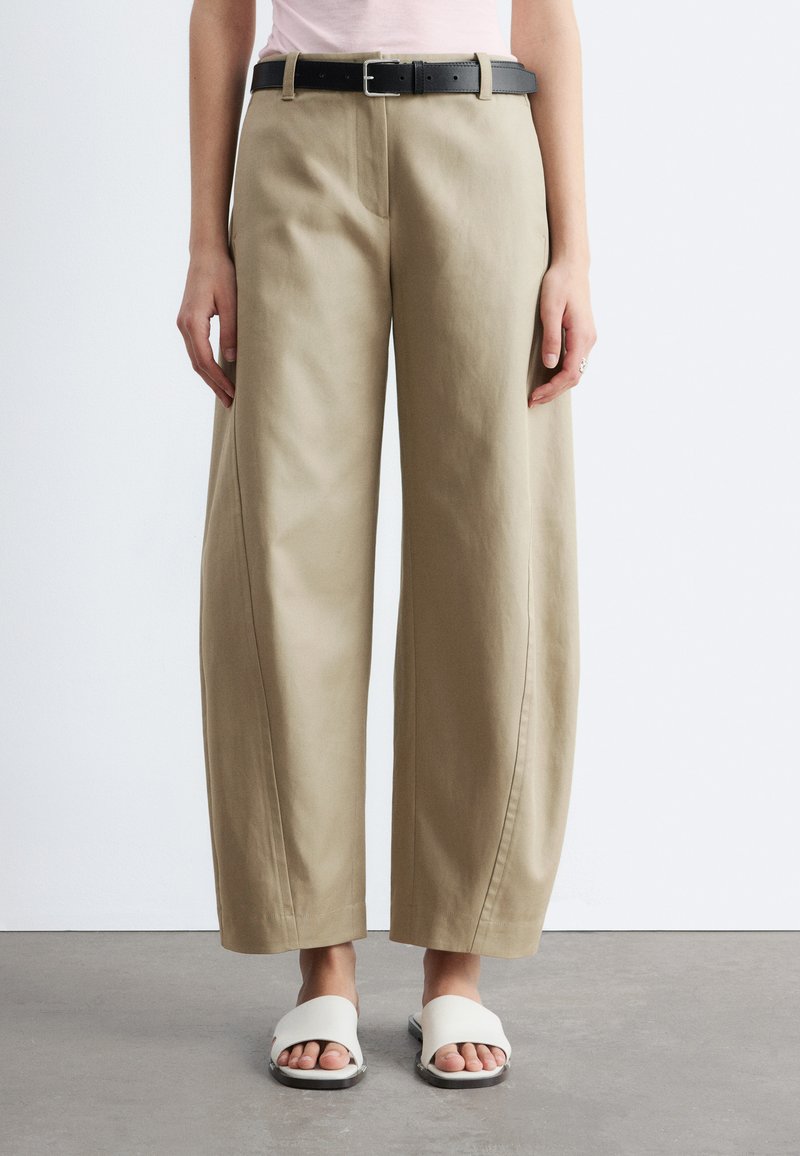 Femme portant un pantalon beige à jambes larges avec une ceinture noire et des sandales blanches à bouts ouverts, debout sur un sol gris contre un mur blanc.