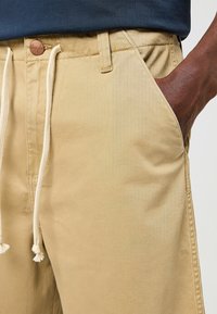 Pantalones cortos de algodón beige con cintura ajustable, bolsillos laterales y un parche de cuero con el logo. La tela tiene una textura suave y un diseño informal.