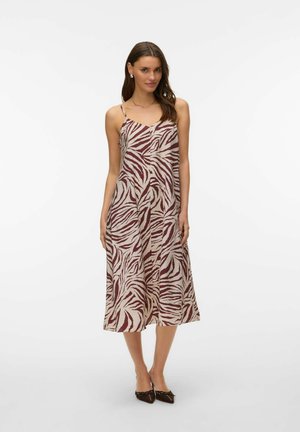 Femme portant une robe beige sans manches, mi-longue, avec des rayures zèbre rouge foncé, debout devant un fond blanc.